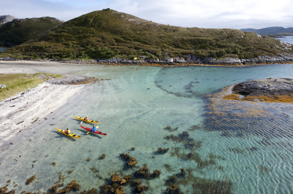 Tre kajakker i blå-grønt vann rett ved en sandstrand på Rødøya på Helgelandskysten. Foto: Terje Rakke.