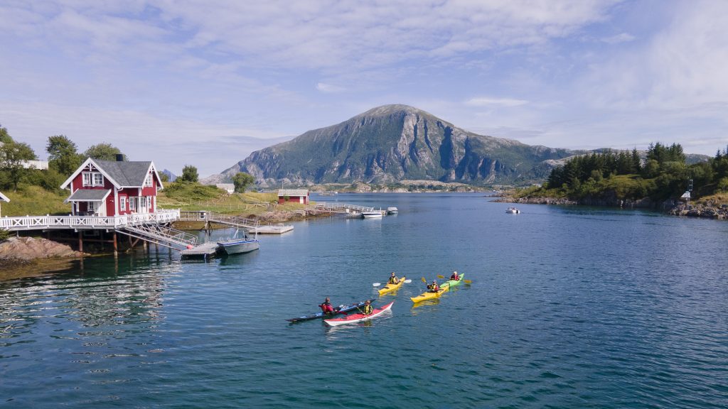 Familie på fire på guidet kajakktur på vei gjennom et sund med guide. I bakgrunnen ser vi båter og Lurøyfjellet, og til venstre et bryggehus.