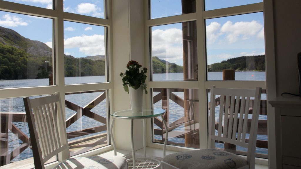 Et rom med to stoler og bord ved to vindusvegger med utsikt til fjord og fjell utenfor veranda i front