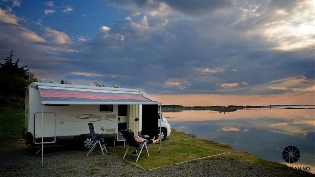 Dame slapper av utenfor campingvogn med utsikt til blikkstille sjø i solnedgangen