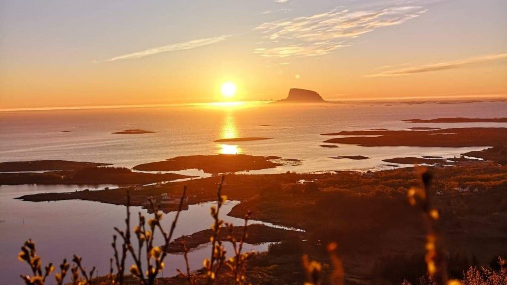 Utsikt fra Dønnesfjellet til havet med solnedgang i horisonten til venstre for Lovund, med flere små øyer, holmer og skjær i havet i mellom