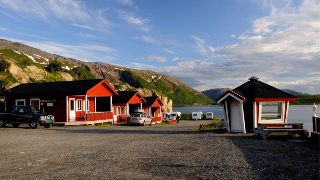 Bildet tatt fra parkeringsplass med utsikt til de røde hyttene og grillhytta i solskinn
