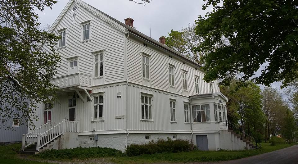 Bildet er tatt utendørs med fokus på det store hvite bygget som har en eldre stil med flere innganger, mange vinduer i det tre-etasjes huset en skyfull dag