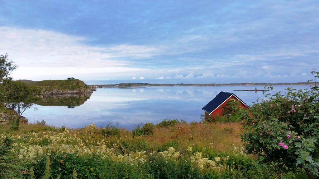 Utsikt fra hage ut til sjøen, med et rødt bygg til høyre i vannkanten og speilblankt hav som reflekterer en himmel full av små lette skyer