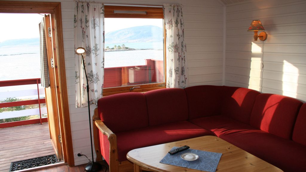 Utsikt fra stue med rød sofa mot stuevindu, og stuebord i tre, med verandadør åpen og utsikt til sjøen