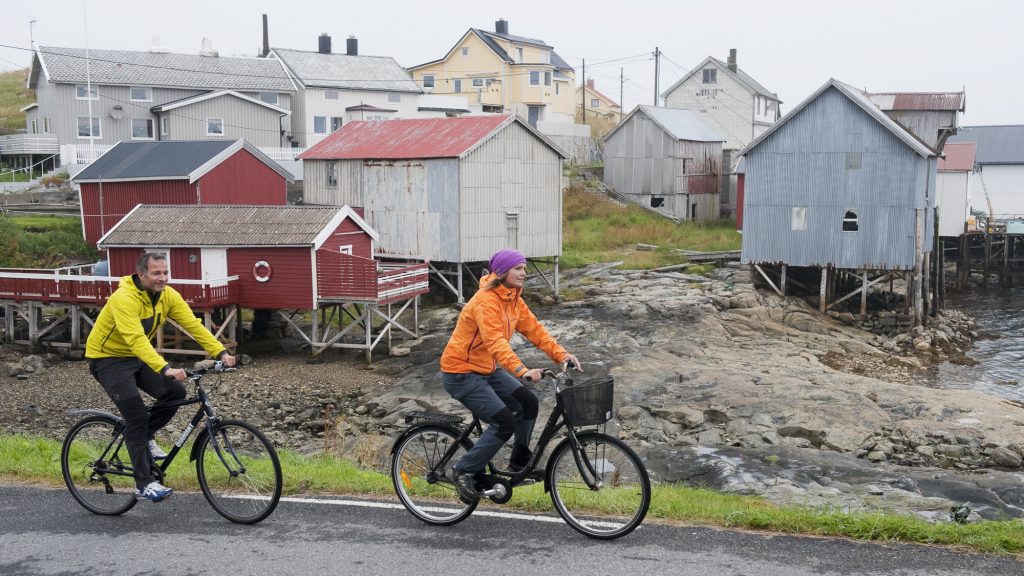 Mann og kvinne i fargerike klær på sykkeltur. I bakgrunnen ser vi vakre, gamle bryggehus