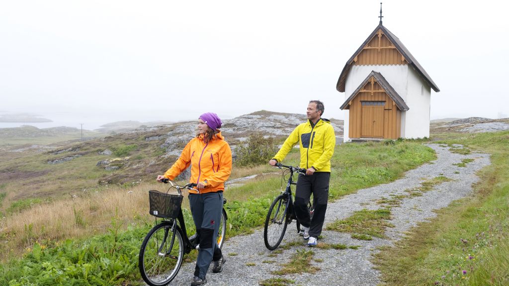 En kvinne og mann i fargerike klær leier hver sin sykkel fra det hvite og tre-brune Petter Dass-kapellet på Træna