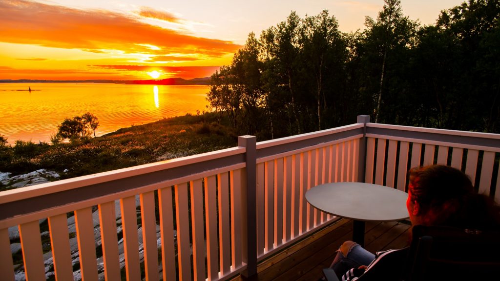 Dame til høyre i bildet sitter på veranda og ser utover havet mot solnedgangen mellom skydotter som speiler seg i havet