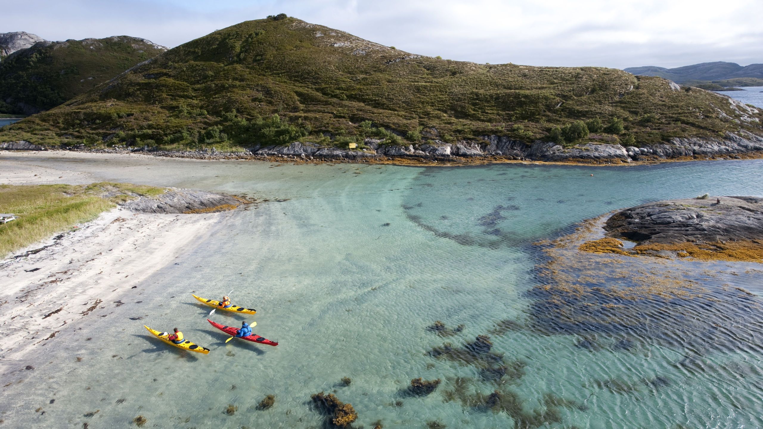 Tre kajakker i blå-grønt vann rett ved en sandstrand på Rødøya på Helgelandskysten. Foto: Terje Rakke.