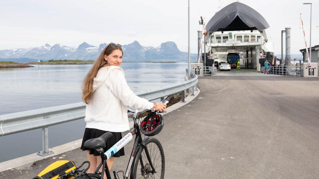 Syklist går med leiesykkel ombord på ferge med utsikt til De Syv Søstre bak fergen. Sykkelen har merke fra Helgeland Reiseliv, hjelm, vannflaskeholder, og bærer med vanntett sykkelveske.