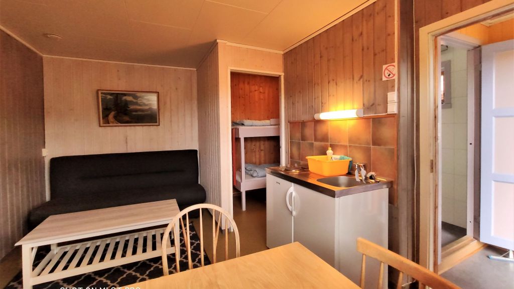 Hytte innvendig sett fra spisebord mot kombinert stue og kjøkken, med åpne dører som viser sovealkove og toalett og utgangsdør.