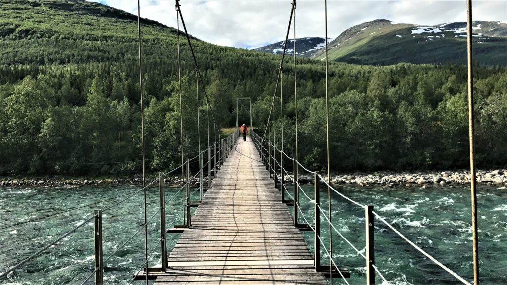 Person på hengebru over ei stor elv. Skog og fjell rundt.