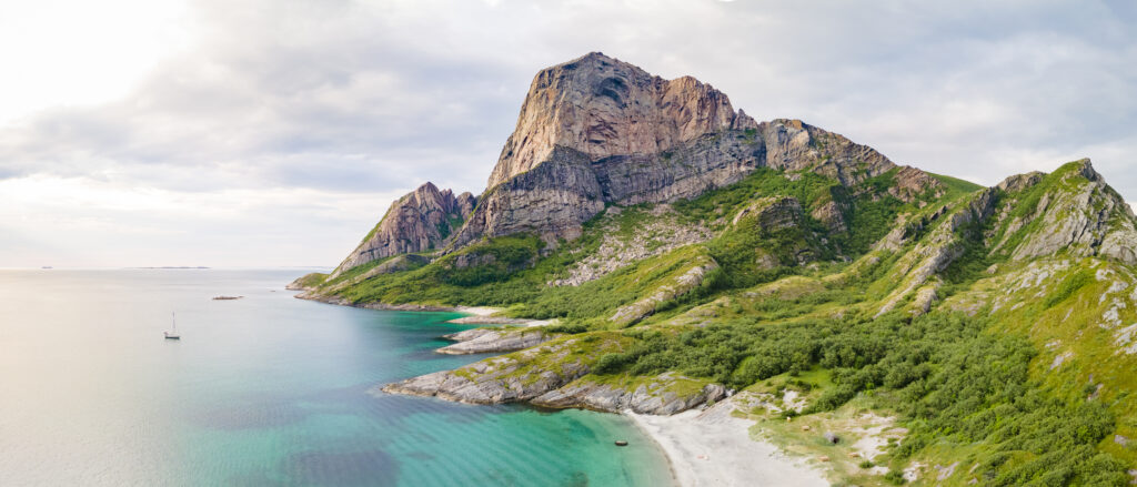 Rødøyløva og Storsadnen på Rødøya sett fra lufta ved solnedgang en finværsdag på sommeren. En seilbåt ligger for anker litt utenfor stranda og fjellet.