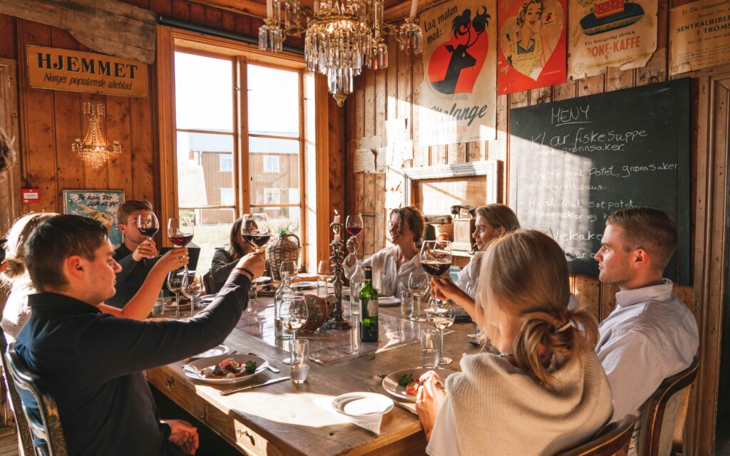 En større gruppe unge menn og kvinner skåler med rødvin ved et koselig middagsbord på restaurant Gammelbutikken på Støtt