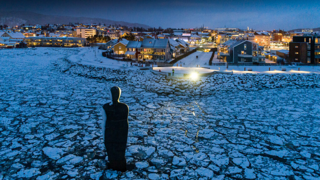 Skulpturen Havmannen foran Moholmen og Mo i Rana en vinterkveld